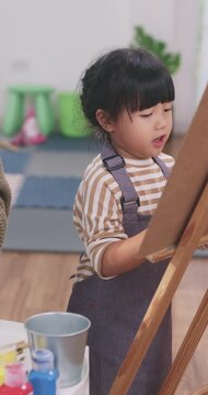 Asian mother teaching her young daughter how to paint on an easel at home.