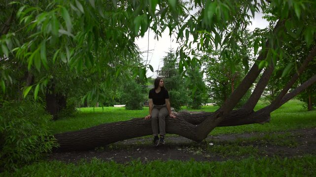 Young woman seated on fallen tree trunk in shaded park, turning head left with calm contemplative posture, hands resting on rough wood, feet dangling above ground