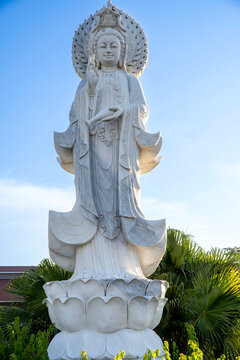 White marble Guanyin bodhisattva statue with halo standing on a lotus base against blue sky.