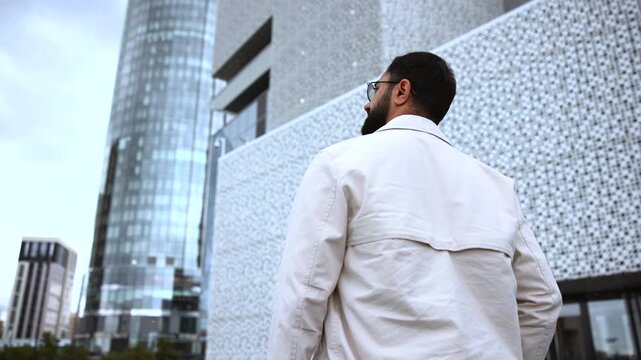 Adult man in light trench coat looking upward at modern glass skyscrapers from urban plaza, seen from rear three-quarter view with cool city atmosphere and clean corporate lines
