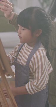 Asian mother teaching her young daughter how to paint on an easel at home.