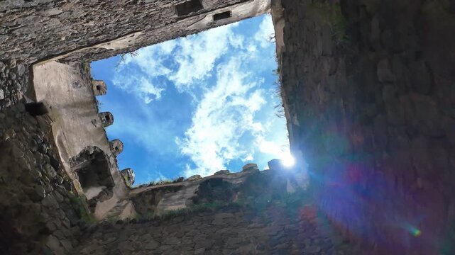 Low-angle perspective looking up at a bright blue sky with white clouds from inside the ruins of an ancient stone castle, with the sun creating a beautiful lens flare effect