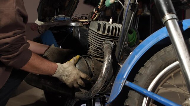 Man wearing protective gloves using tools to repair an old, classic motorcycle engine and components, performing maintenance in a workshop environment