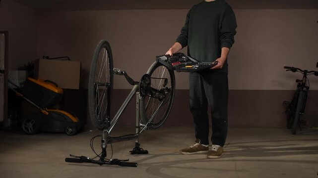 Man in a garage standing next to an upside down bicycle on a stand, holding an open tool kit, preparing for maintenance or repair work on the bike