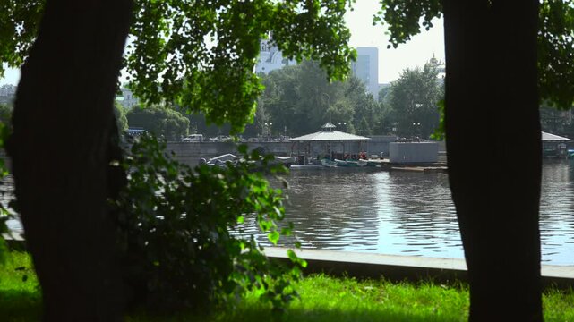 Shaded riverside park viewpoint framing marina pavilion, moored rowboats and reflective water ripples beneath leafy canopy with strong midday backlight, with river, marina, pavilion, rowboat detail