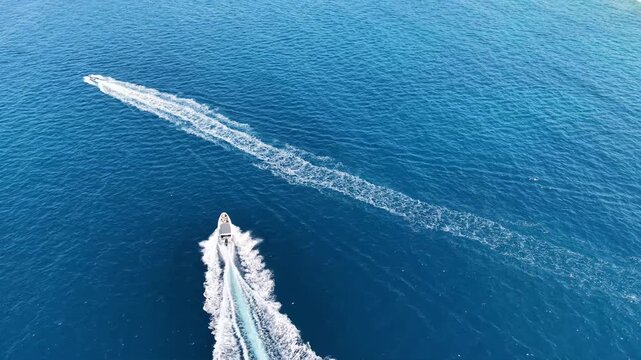 Drone footage: motorboats leave white foamy wakes on the clear, dark blue water. A picturesque summer seascape featuring a green coastline, a sandy beach, and resort infrastructure set against a backd