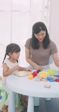 Asian mother and daughter playing kitchen pretend game, cutting toy vegetables on a wooden board.