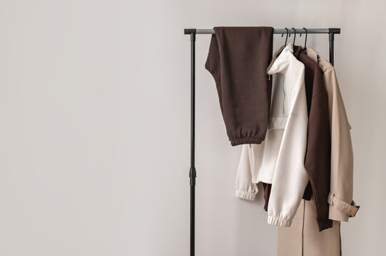 Elegant minimalist aesthetic woman clothing capsule, autumn wardrobe in brown and beige colors, jersey sweaters, pants and trench coat on clothing rack, empty neutral white wall background copy space