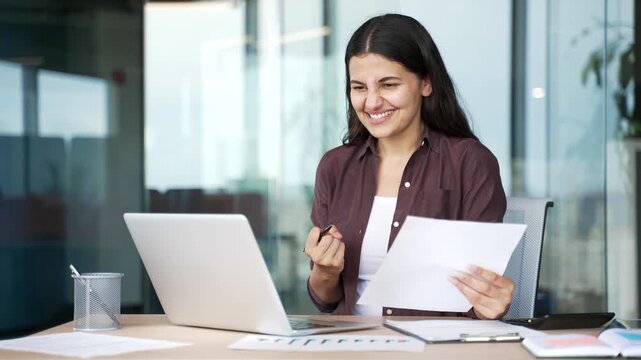 Smiling young businesswoman reviewing financial documents on laptop and satisfied with results sitting in business office. Happy female financier rejoices at positive indicators, celebrates success.