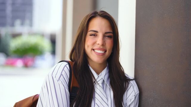 A woman stands outside a building in a city. She has long hair and is wearing a striped shirt with a backpack. The sun is shining and people can be seen in the background.