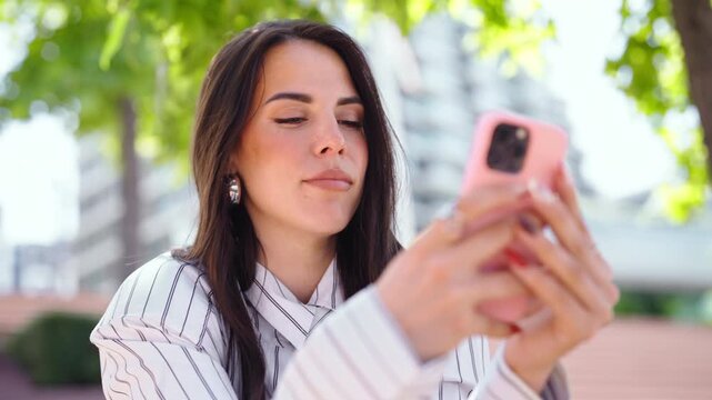 A woman sits on a park bench in a city. She is focused on her smartphone while enjoying the shade from trees. People walk by in the background.