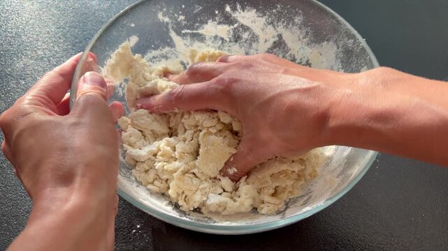 a person's hands kneading a shaggy, raw dough mixture inside a clear glass bowl.