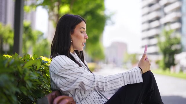 A woman sits on a sidewalk in the city and uses a tablet. She smiles as she looks at the screen. Tall buildings and trees surround her. It is a sunny day.
