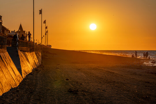 Saint-Aubin-Sur-Mer, France - 07 27 2026: View the jetty, sand dune, national flags, people, the sea and the sun at golden hour