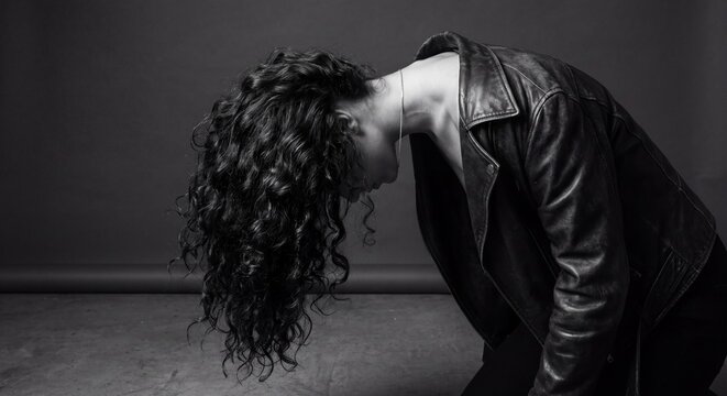 Dramatic black and white studio portrait of a woman with long curly hair bending forward wearing a leather jacket