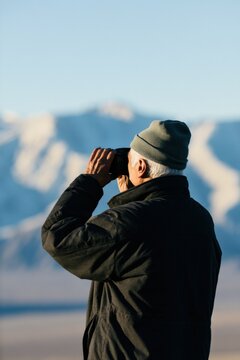 Senior man looking through binoculars in mountain landscape background for travel and outdoor adventure banner and wallpaper