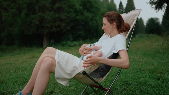 Young mother sits in a comfortable folding chair outdoors, breastfeeding her newborn baby. She tenderly holds the infant, creating a loving connection amidst the natural scenery of a green park