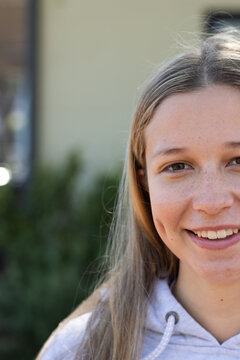 Teenage female smiling, facing camera while standing by house wall with shrubs wearing gray hoodie