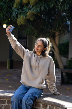 African American teen female sitting on brick-wall wearing hoodie taking phone selfie in courtyard