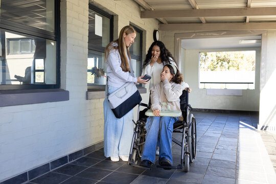 Diverse female teens showing phone to seated student in manual wheelchair with lap tray on corridor