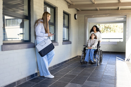 Diverse women group walking in covered corridor, pushing child in wheelchair other checking phone
