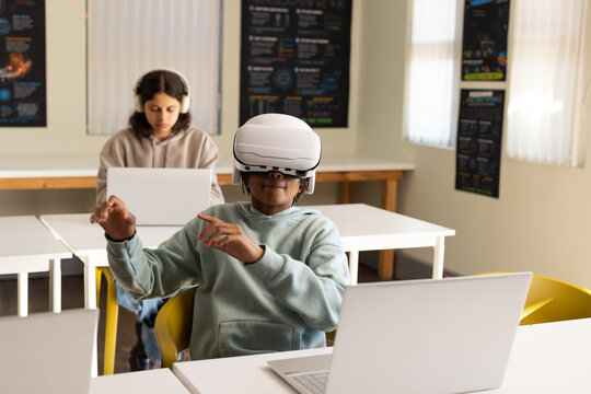 African American youth students wearing hoodies using white VR headset and laptops in class