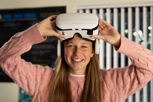 Youth female holding VR headset above head at workstation with monitor showing code in pink sweater