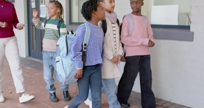 Diverse female pupils walking school path with teacher gesturing, girls stopping to view bulletin
