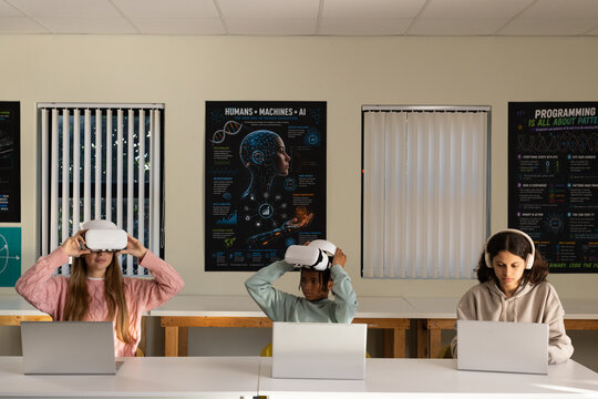 Diverse teenage students using white VR headsets and working on silver laptops at tech lab