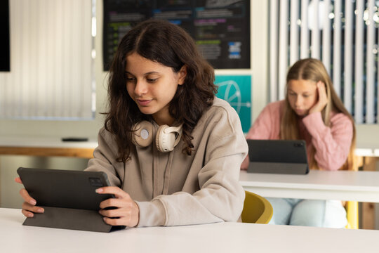 Diverse female peers using tablets and studying in classroom, wearing beige hoodie with headphones