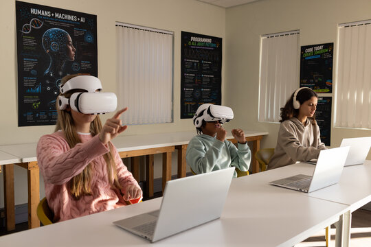 Teenage students wearing pink sweater, beige hoodie using white VR headsets and laptops in tech lab
