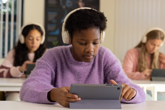 Diverse teen girls in class using tablets and wearing white headphones, center in purple sweater