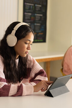 Diverse teenage female peers sitting at study table wearing white headset using tablet in gray case