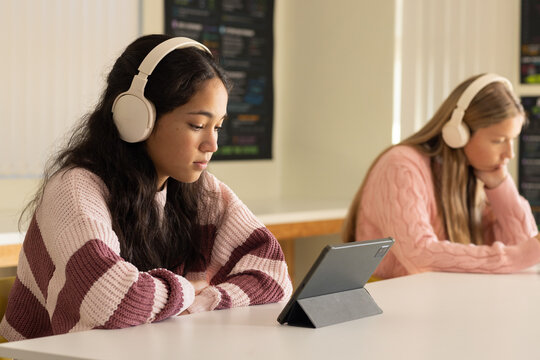 Diverse teenage girls studying in class at table using tablet, wearing headphones, copy space