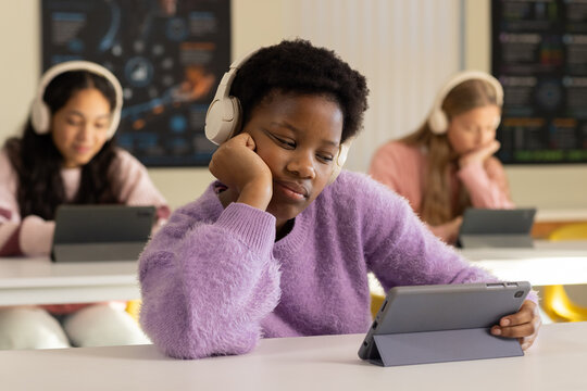 Diverse teenage girls in classroom using tablets, front girl wearing purple sweater with headphones