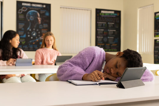 Teenage girls in class leaning at desks, one in lavender sweater writing in notebook using tablet