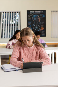 Three female youths at white desks in classroom, foreground wearing pink sweater writing on tablet
