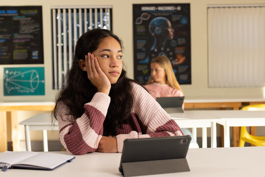Diverse female teens in class left teen wearing striped knit gazing right using tablet notebook