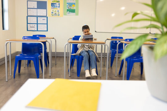 African American child girl sitting at front-row desk using tablet in classroom with blue chairs