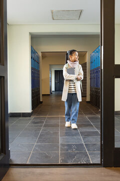 Teenage female student wearing coat and backpack, standing in school hall with binder and lockers