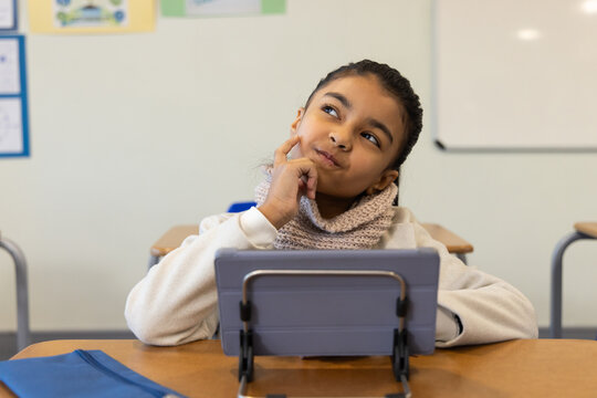 Preteen female student sitting at school desk behind tablet on stand, touching cheek, blue folder