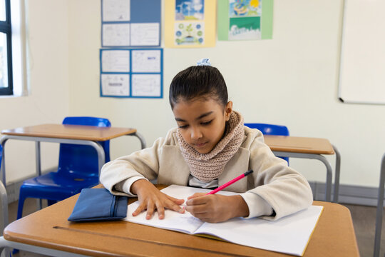 Indian school-age girl writing in open notebook in classroom with pink pencil, in beige coat, scarf