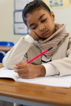 Female school-age child sitting at desk in class writing in book with pink pencil wearing scarf