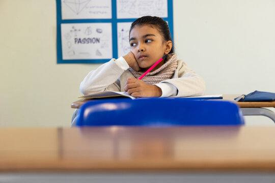 Asian female child sitting at school desk, wearing scarf, holding pink pencil above notebook