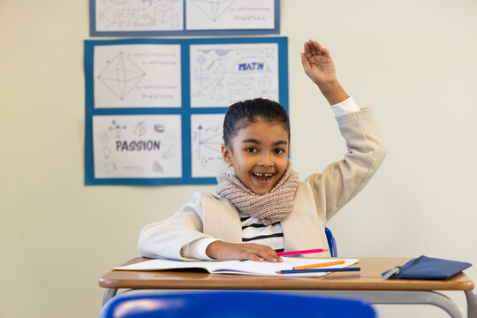 African American child wearing scarf raising hand sitting at desk with notebook and colored pencils