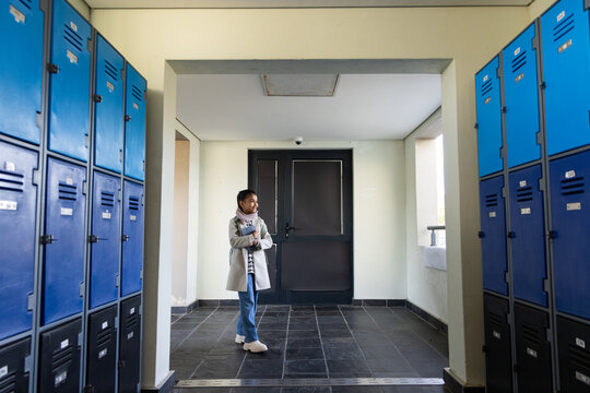 African youth woman standing in tiled hall holding notes and phone looking right by blue lockers