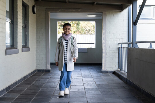 Teenage African American girl walking down tiled corridor toward camera wearing coat scarf backpack