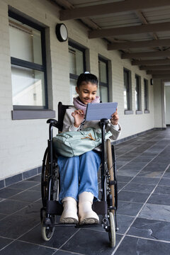 African American teenage female sitting in wheelchair using tablet with backpack on lap in hall