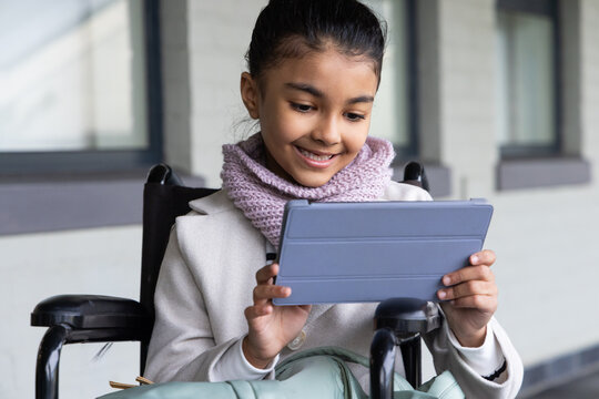 African female youth sitting in wheelchair holding tablet folio, smiling with scarf beside building