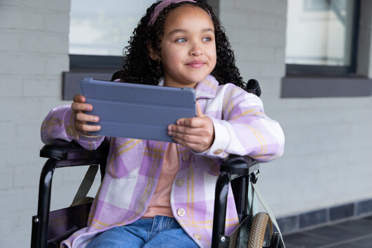 Youth African American female sitting in wheelchair under walkway holding tablet in plaid jacket